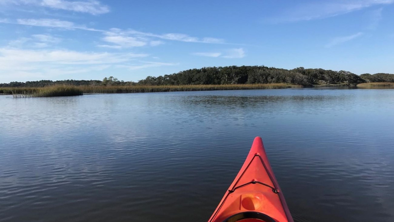 Front of kayak surrounded by water, overlooking salt marsh and vegetated island with clear sky above