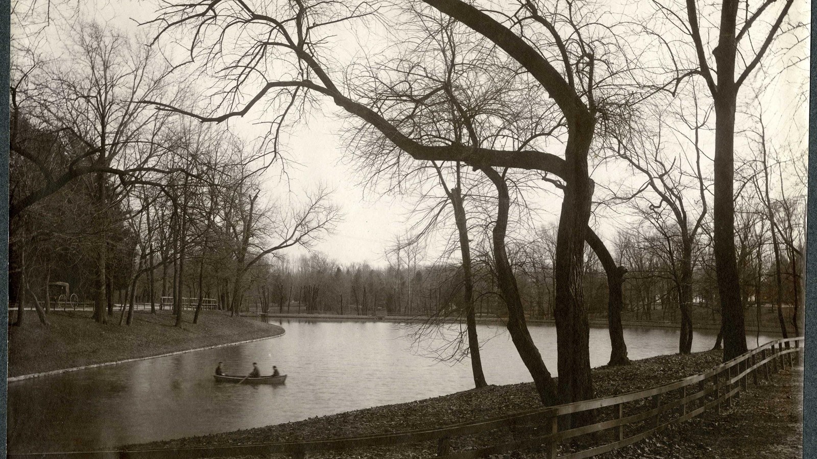 Black and white of body of water with people on canoe and trees and grass on side of water