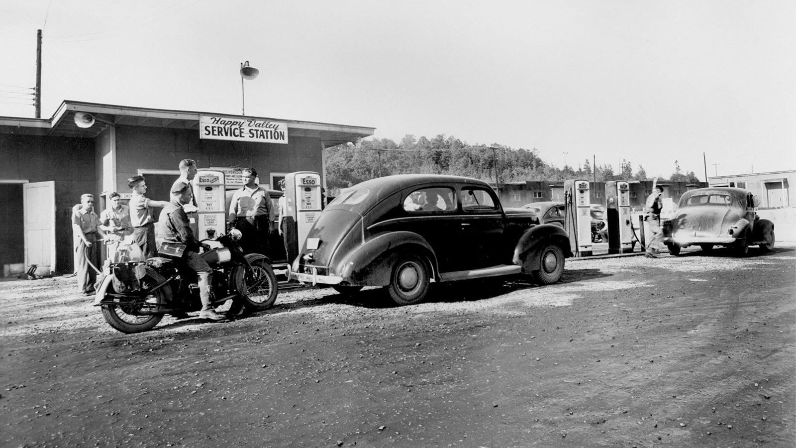A small service station surrounded by people, cars, and motorcycles