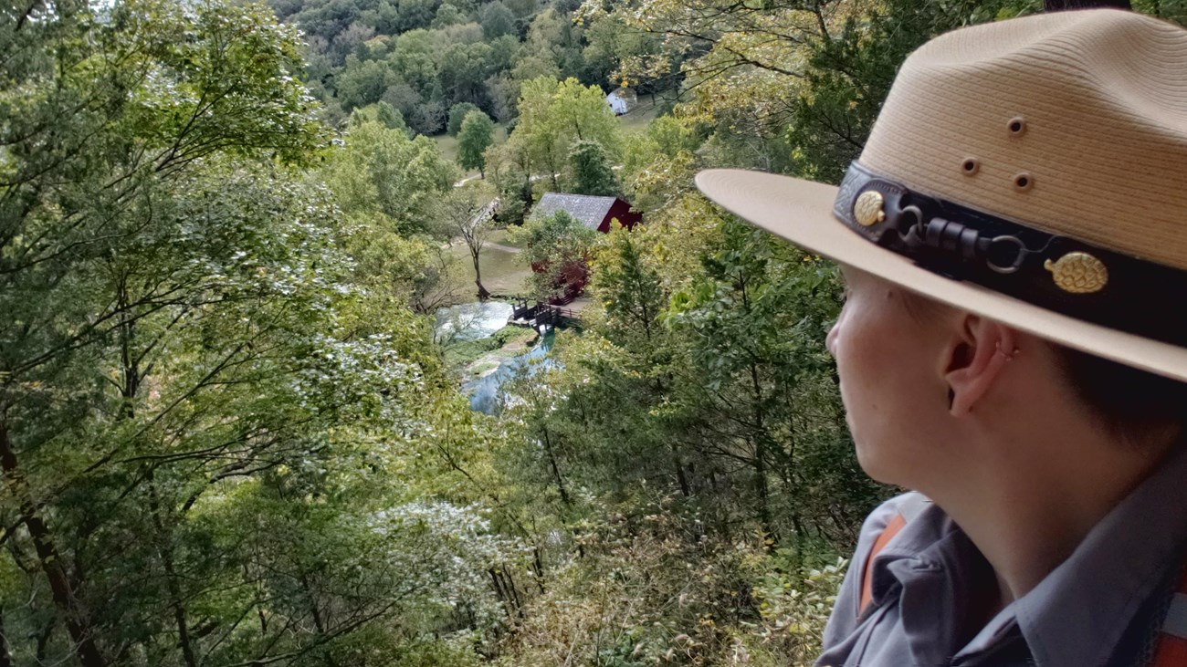 a female ranger in flat hat and uniform on overlook with red mill and blue spring below
