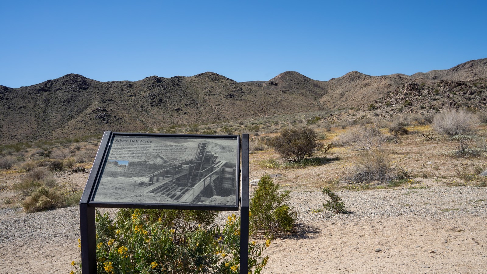 A wayside next to a desert landscape. Mine remnants can be seen on distant rocky hill.