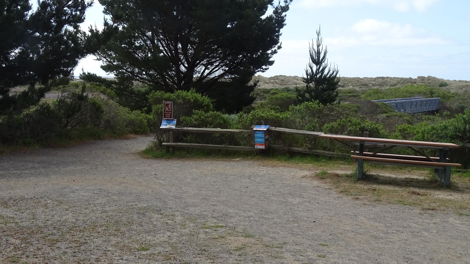 A path passes between some trees on the left and a picnic table and a split-rail fence on the right.