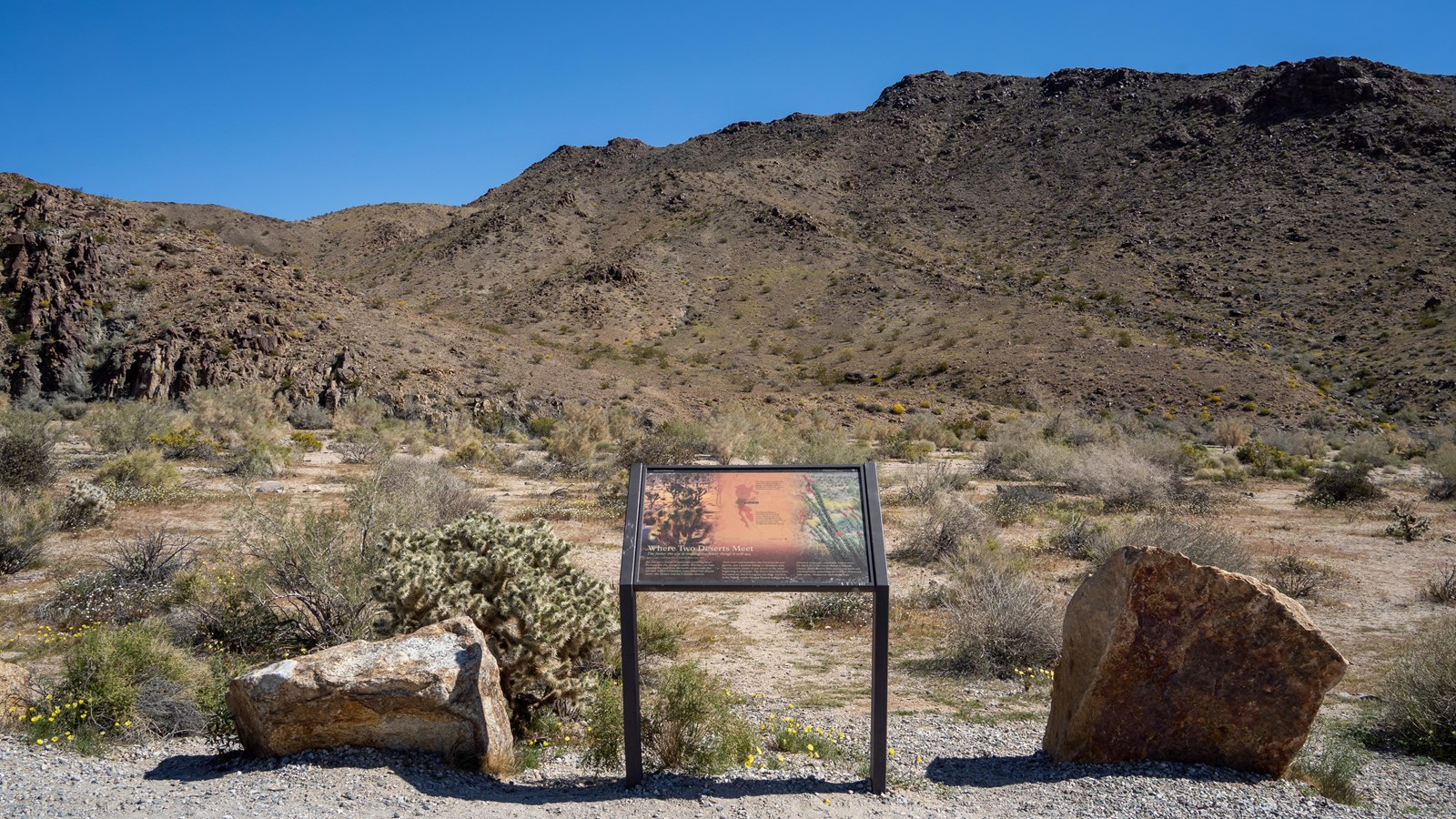 A wayside sign stands at the edge of a pullout between two rocks. Rocky hills in the background.