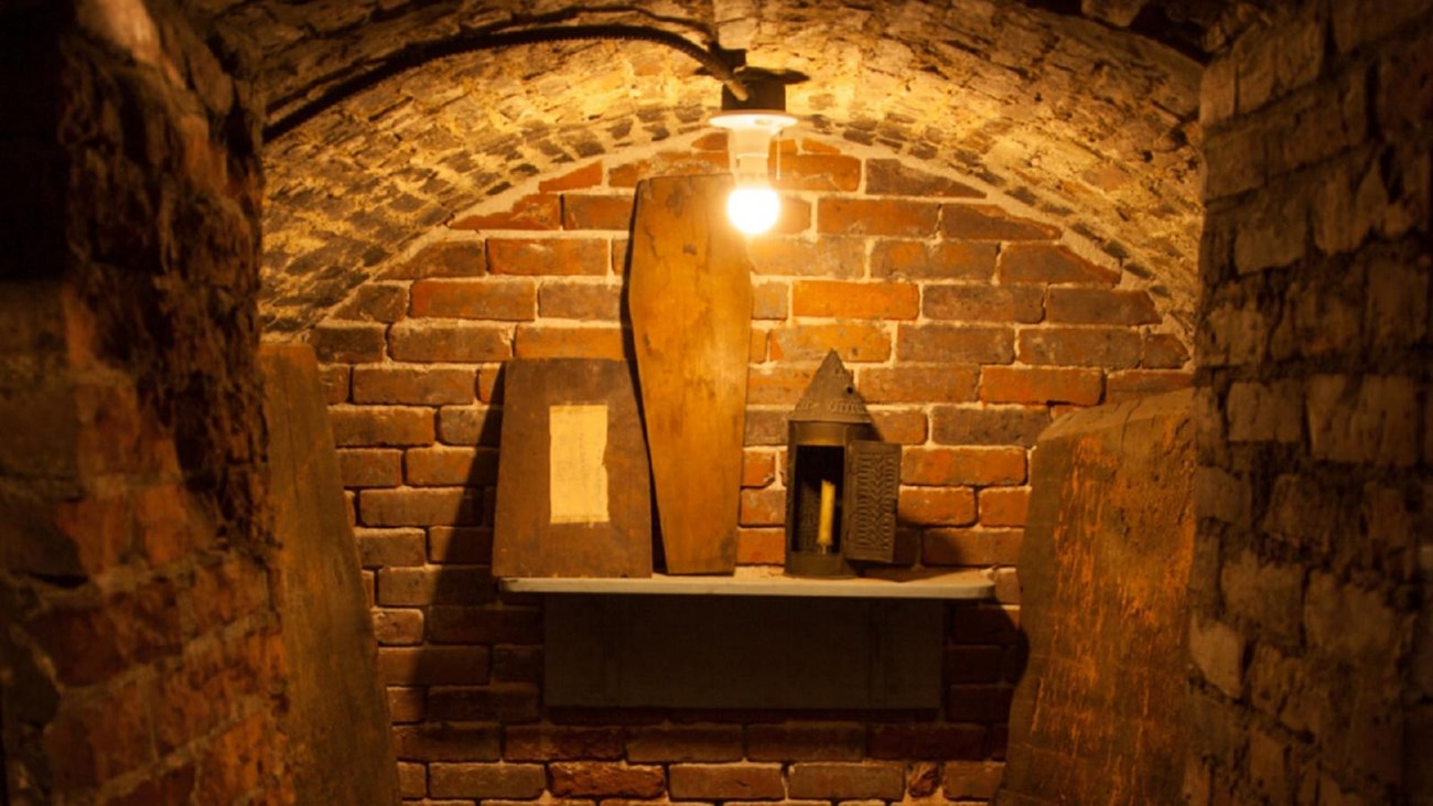 Inside a small red-brick crypt with a shelf that has a couple boxes and a coffin shaped box.