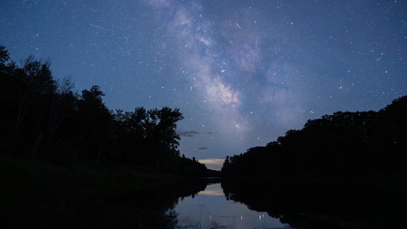 The Milky Way and many stars illuminate the night sky over the river.