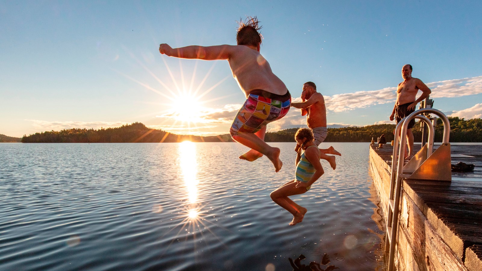 Three people jump off a dock into water as one person watched while the sun sets.