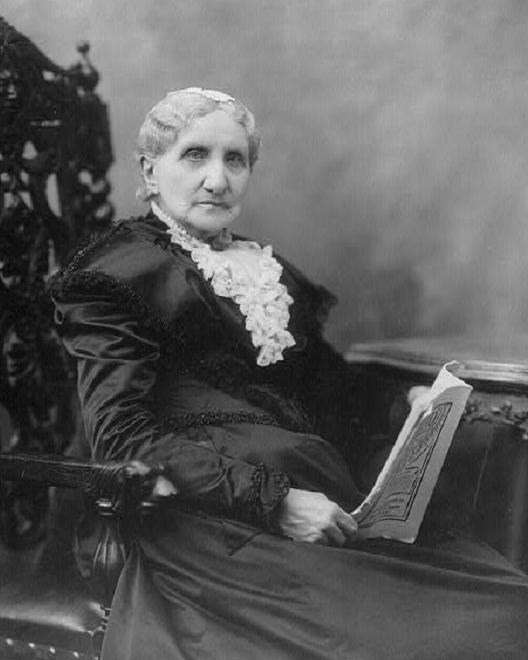 An old woman with white hair in a black dress sitting in a large chair. 