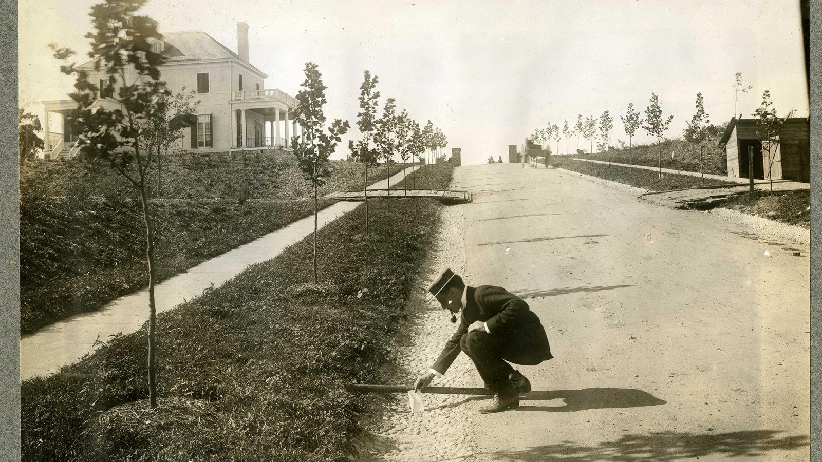 Black and white of dirt road with man in suit bending down. Road lined with grass and trees
