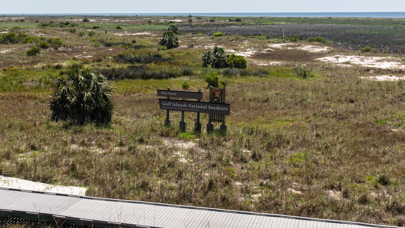 A sign welcoming visitors to Ship Island stands behind a boardwalk in the middle of a grassy plain.