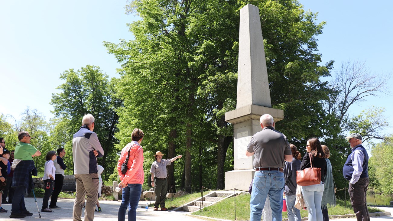 A park ranger stands before a stone monument pointing and talking to a crowd of visitors