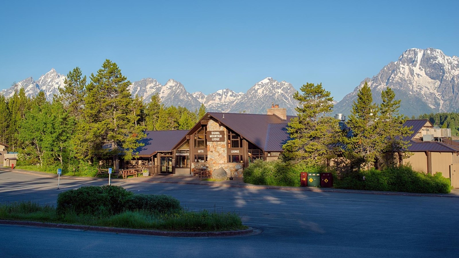 A wide angle image of a wooden lodge with trees and mountains with a blue sky.