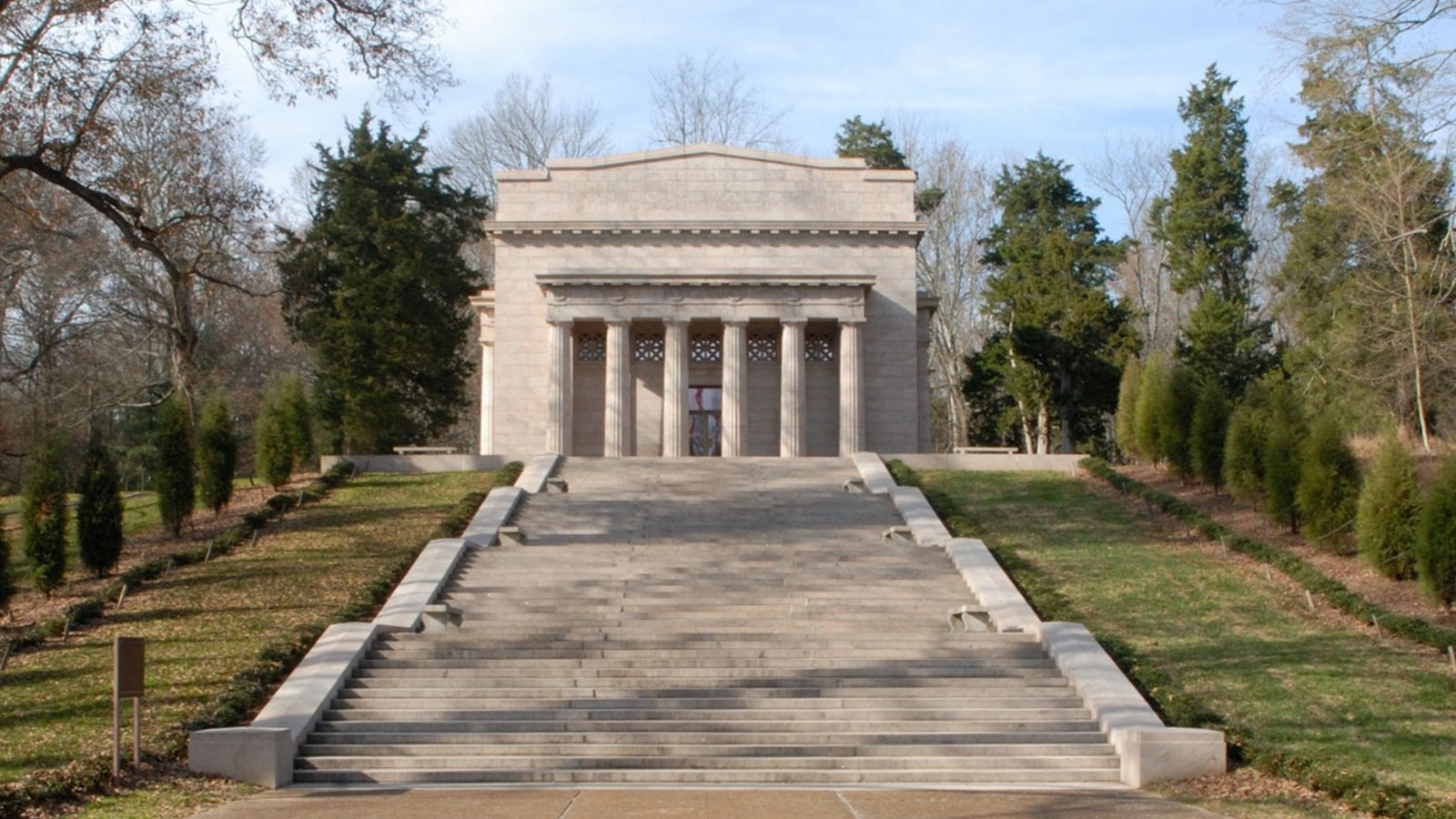 Stone structure that is greyish-pink with columns on top of a hill stone with steps leading up. 