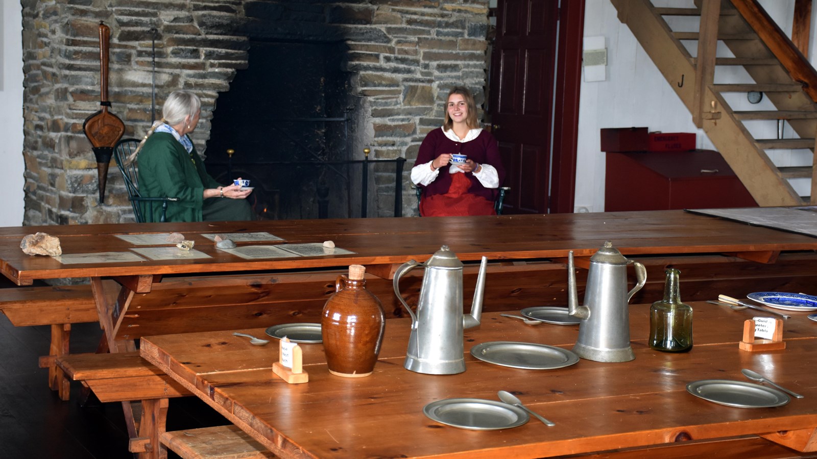 Two people wearing historic clothing, seated before a fireplace, holding tea cups.