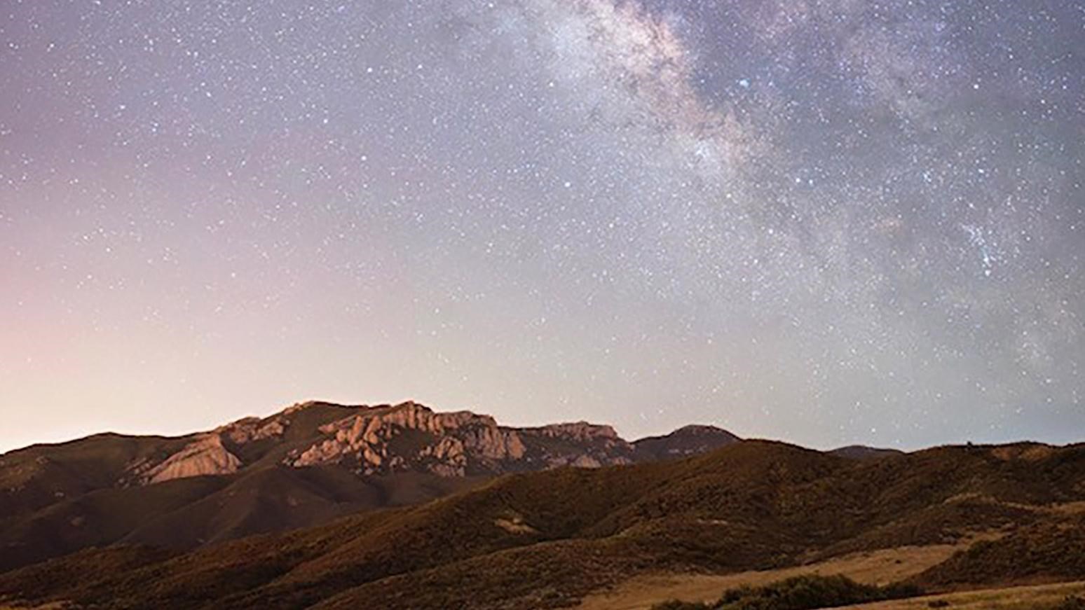 The Milky Way among stars in a night sky over a rocky mountain.