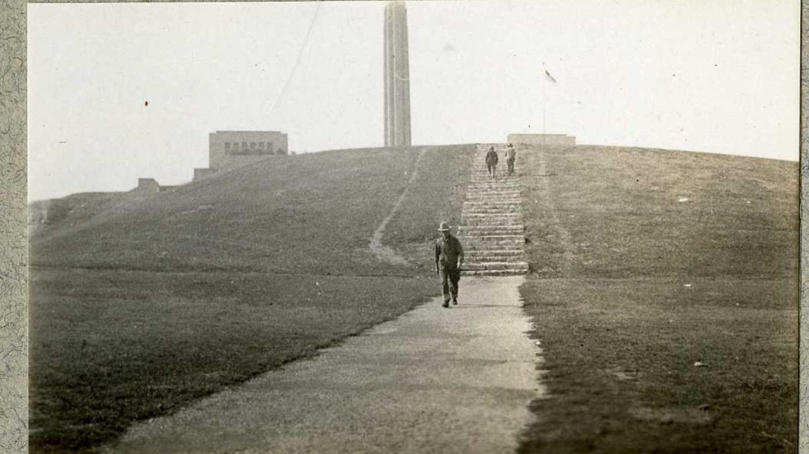 Black and white of grassy hill wit stairs in it and pillar up top with people walking on path