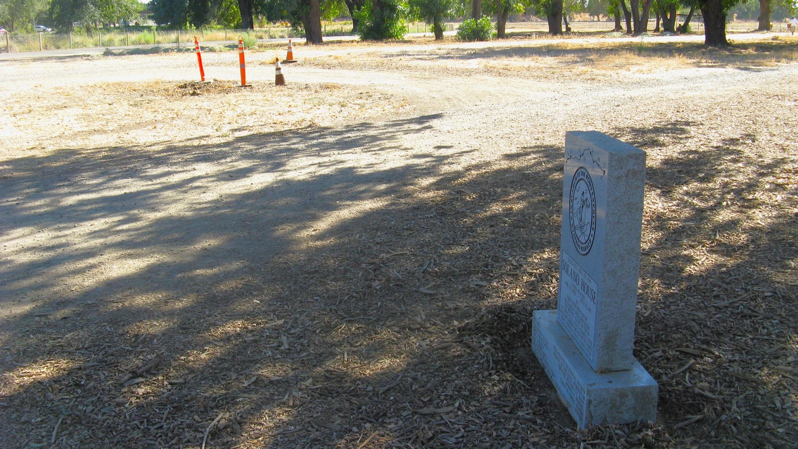 A small granite monument stands among wood chips in a shaded area along a bike path. 