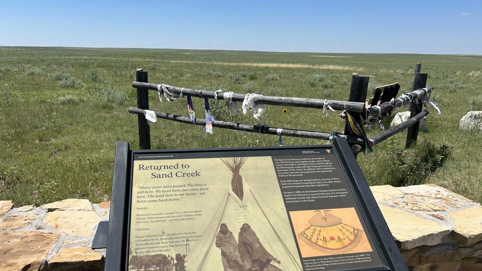 An exhibit stands before a small wooden fence with prayer ribbons and an open field beyond. 