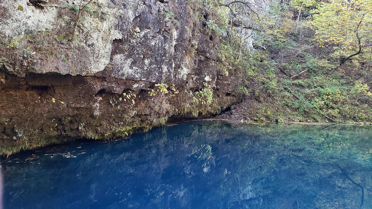 a deep dark blue spring at the bottom of a cliff of gray rock