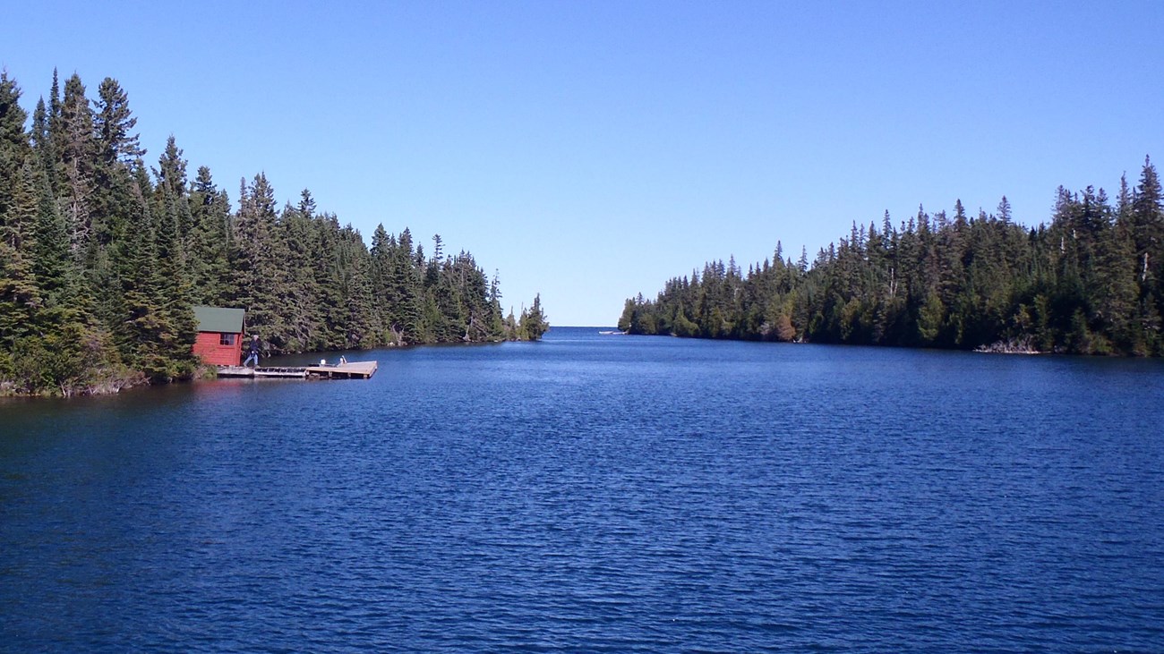 A red historic summer cottage mail room and dock can be seen along the conifer lined Tobin Harbor. 