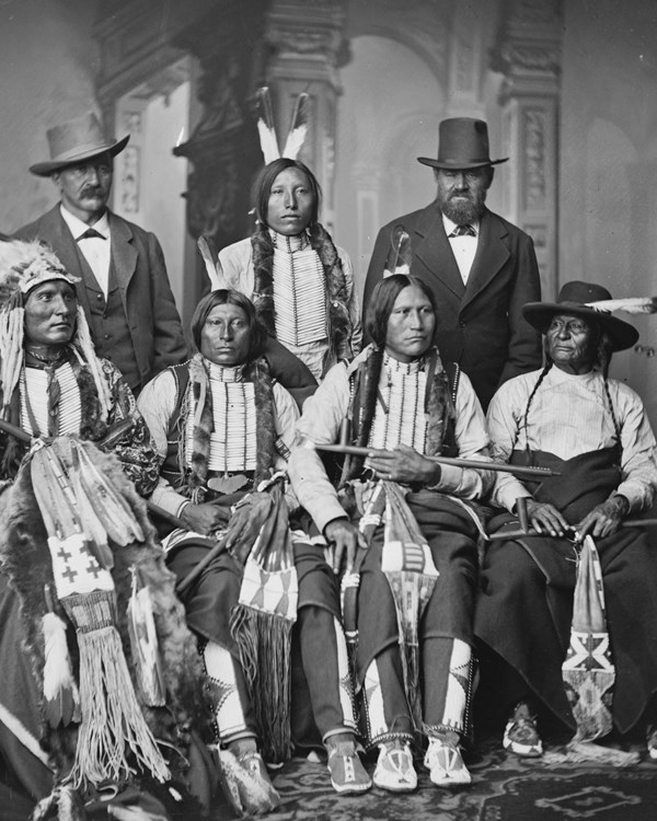 Portrait of five Arapaho nation men in traditional clothing and two men in suits behind them.