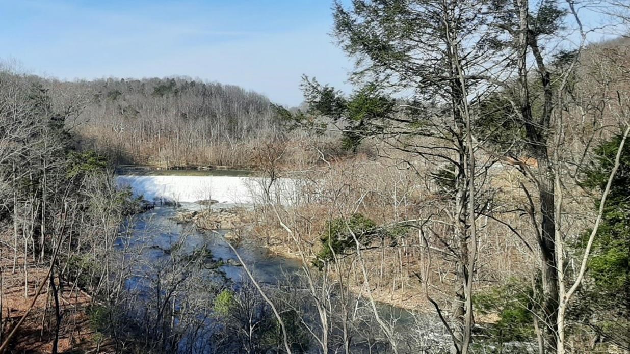 A low, white dam on a river surrounded by winter-bare forested hills