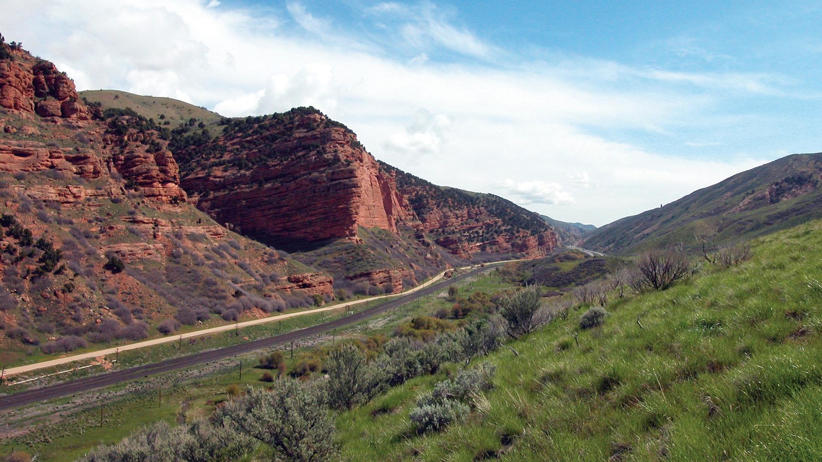 A paved highway leads away through a canyon lined with red, rocky cliffs, and grassy hills.