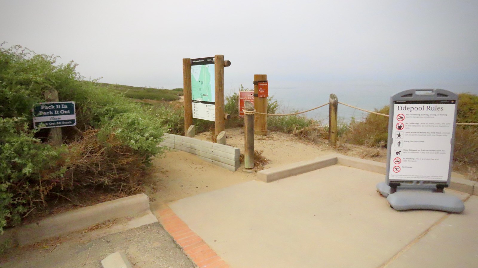 Several signs mark the end of the dirt trail. Bushes are on left and the ocean is in the distance.
