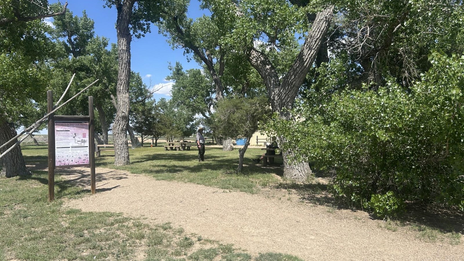 A park ranger speaks to visitors in a grassy area with trees, trails, signs, and picnic tables. 