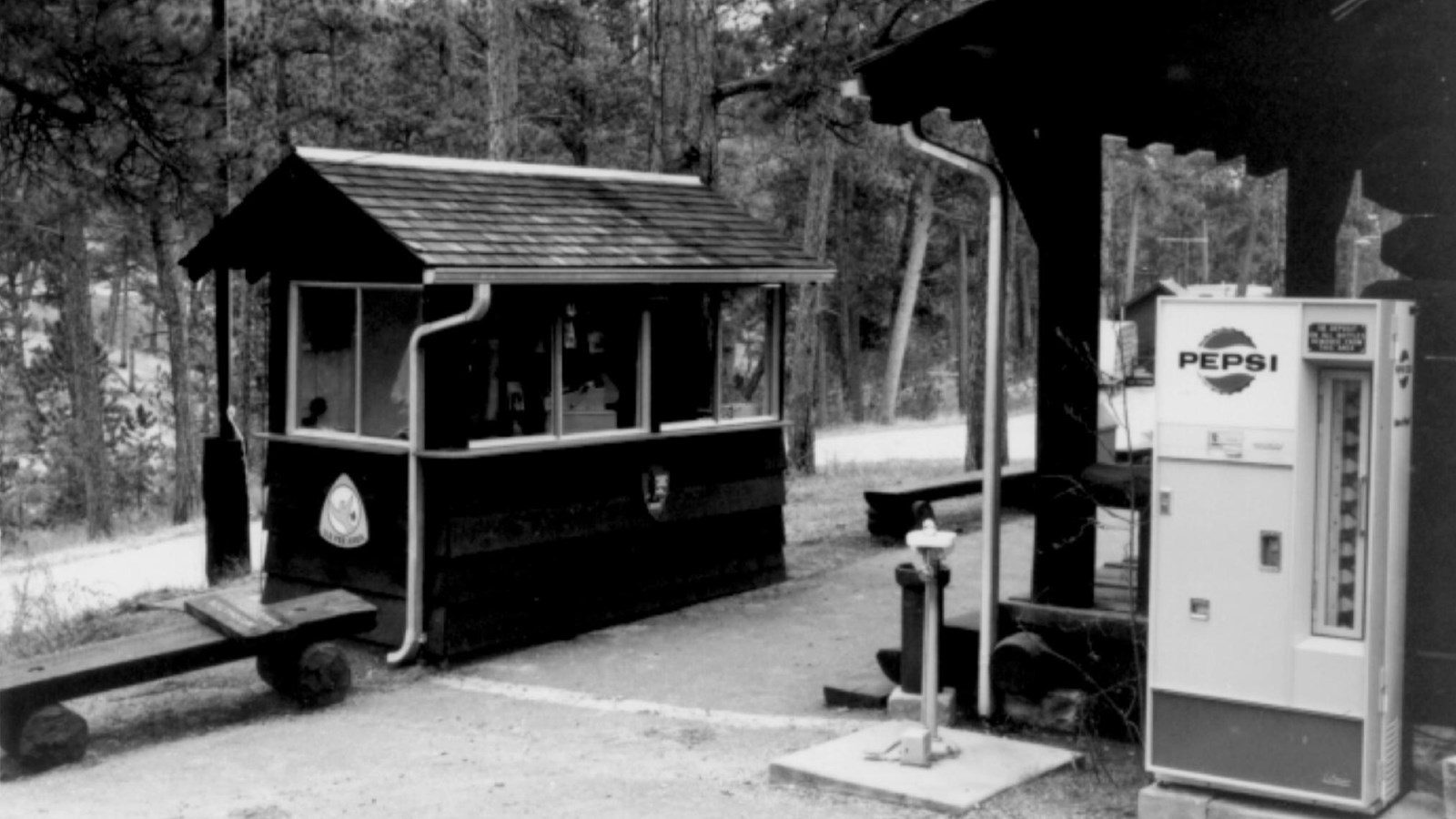 A black-and-white photo of a small fee kiosk, Pepsi machine, and part of the Historic Cabin’s front