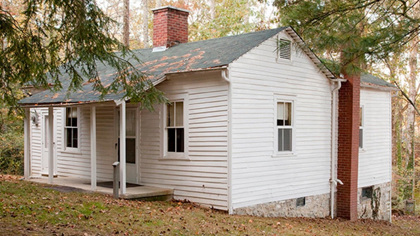 Small white wooden house has two doors opening onto a front porch, with brick chimney.