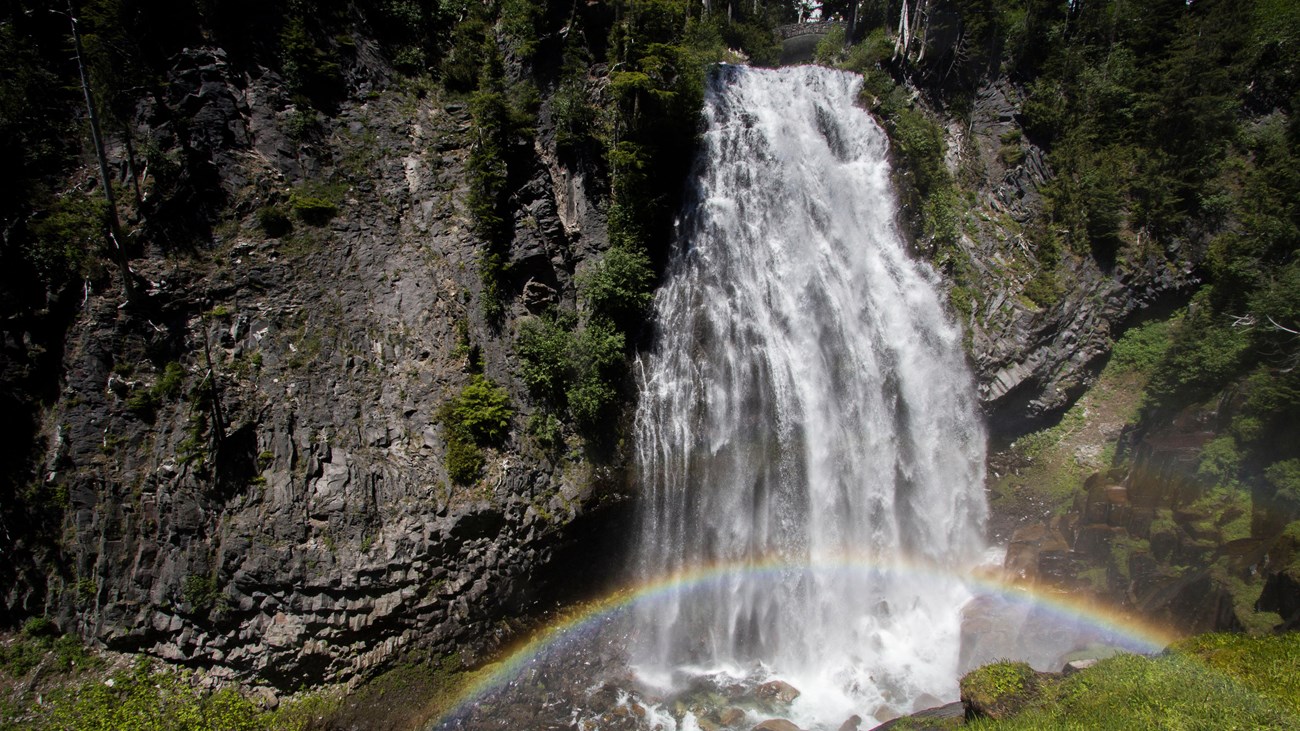 A waterfall cascades off a cliff with a rainbow arching through the mist of the waterfall. 