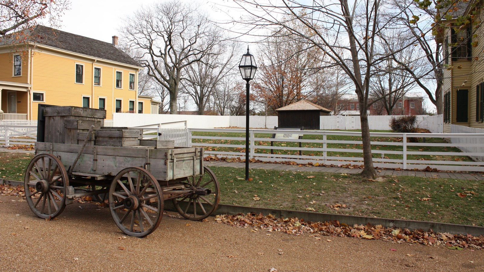 Empty grass lot surrounded by white fence with old wooden wagon/cart in front on street