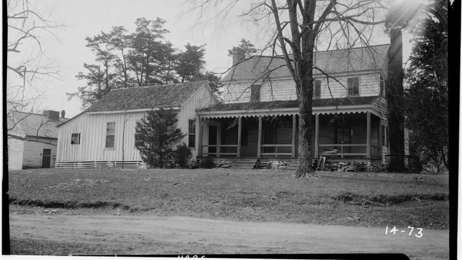 Black and white photo of a two story farmhouse with a covered porch and a brick chimney