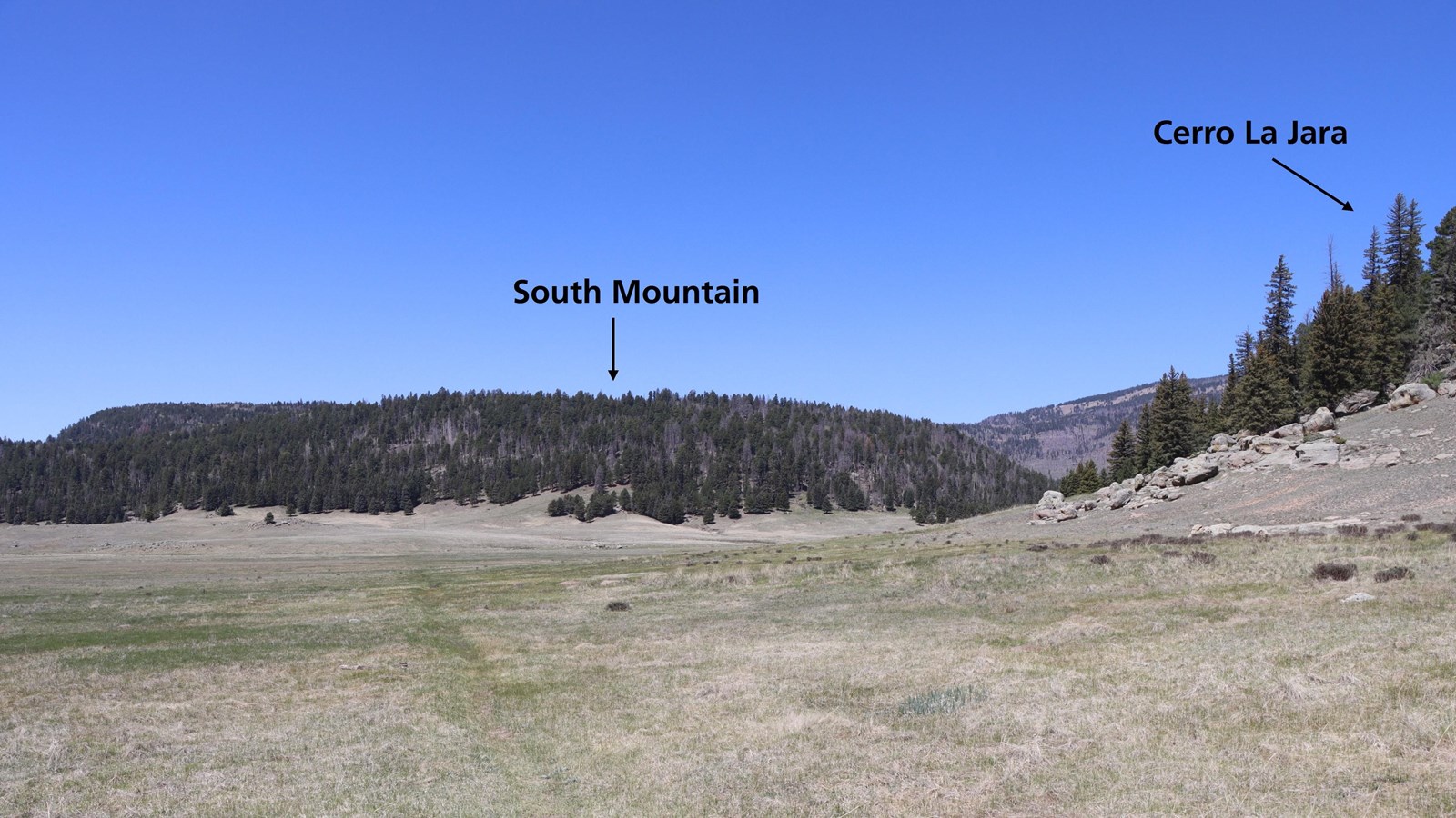 A narrow, dirt path wraps around a lava dome. In front of us is a flat-topped, forested mountain.