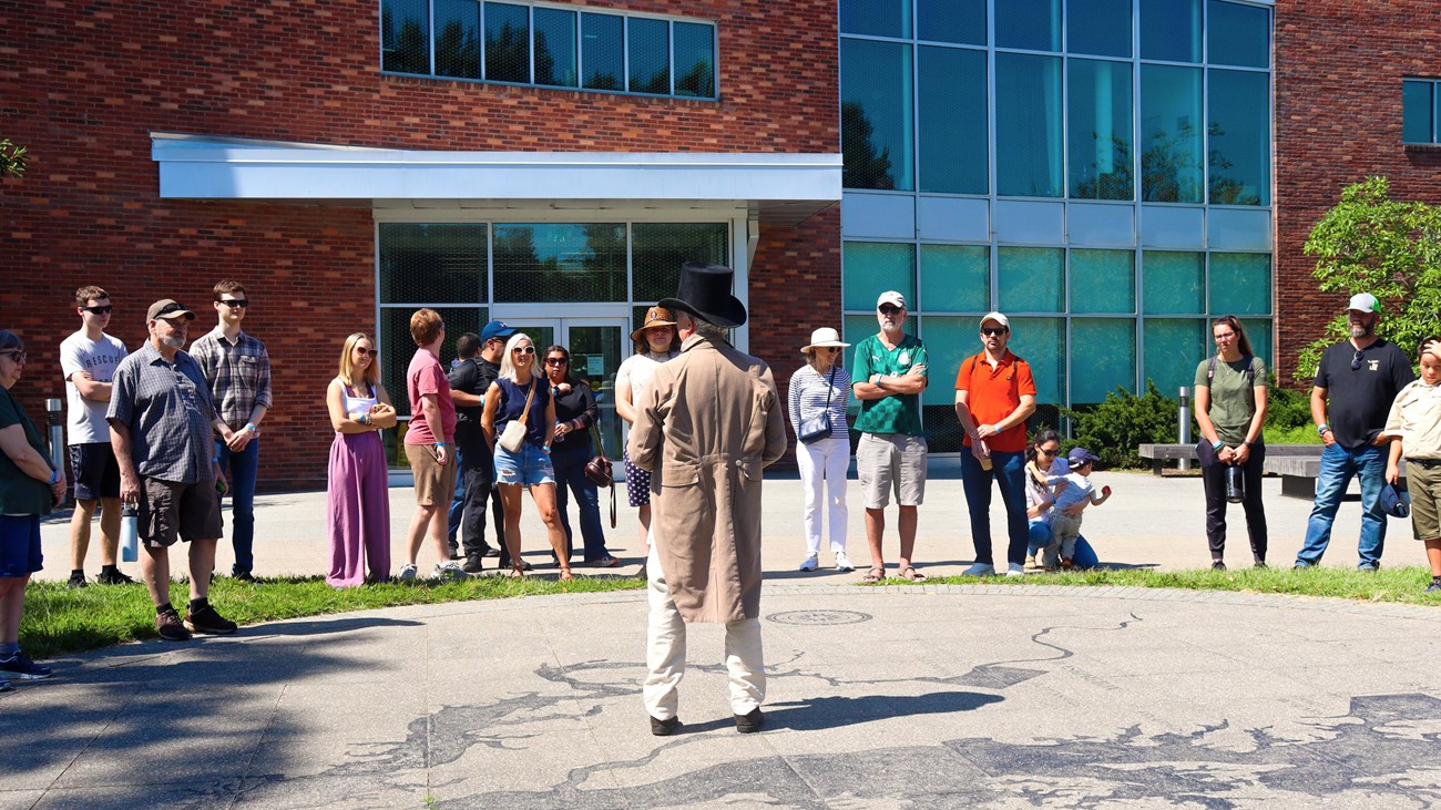 Ranger in living history garb speaks to a semicircle of people outside of the visitor center.