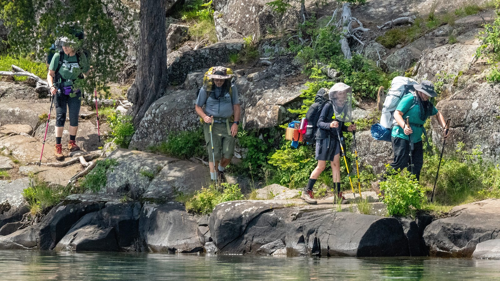 Four people with large backpacks and hiking poles hike on a rocky shoreline.