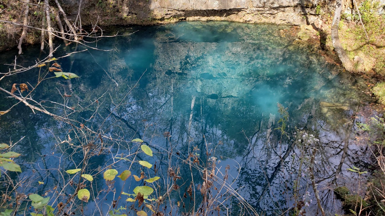 a deep blue spring is at the bottom of a cliff face of grey rock