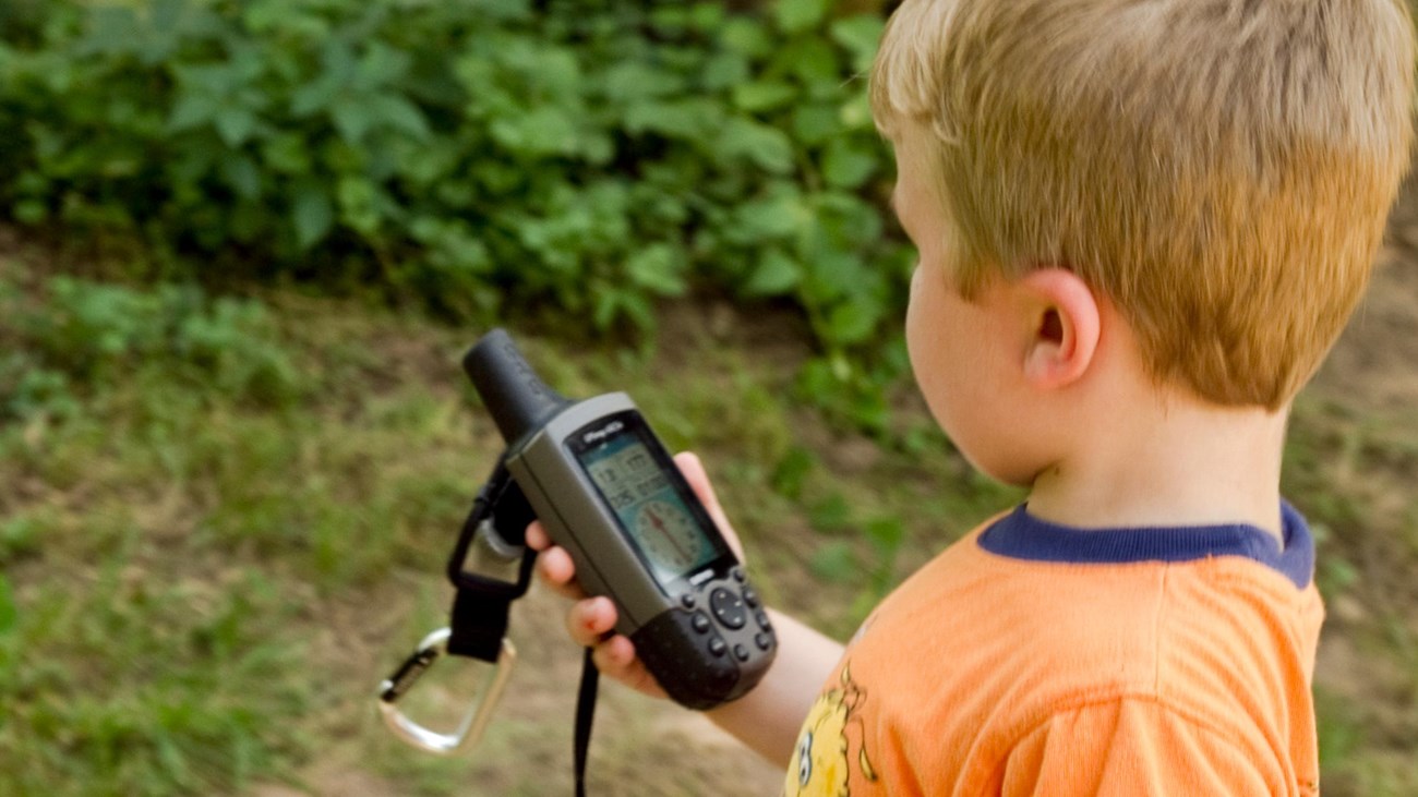 A boy wearing an orange shirt holds a GPS device in his hand