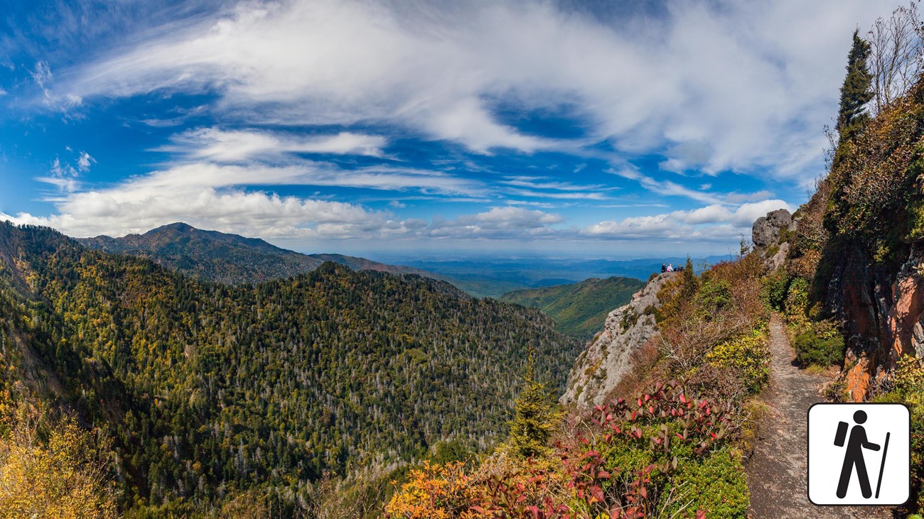 A narrow trail with sweeping views to the left on a partly cloudy day. A hiker icon in corner.