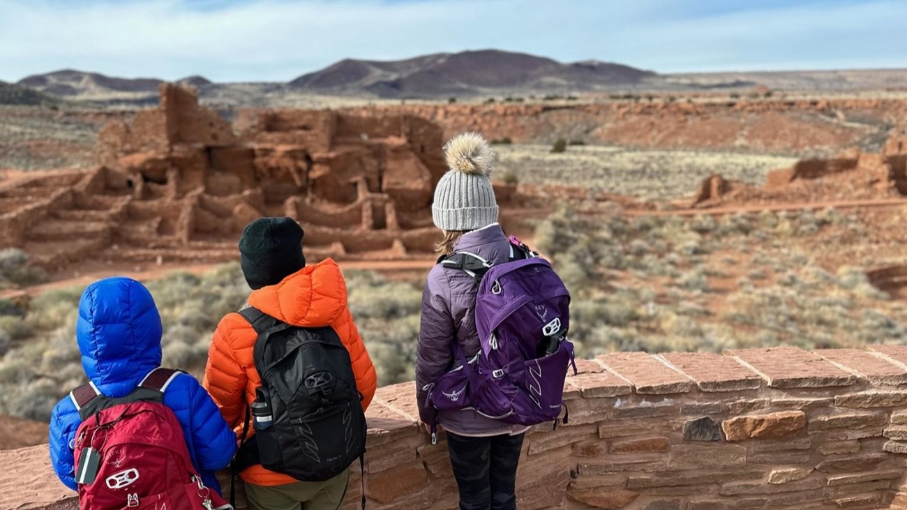 Three Jr. Rangers standing at the Wupatki trail overlook viewing Wupatki Pueblo.