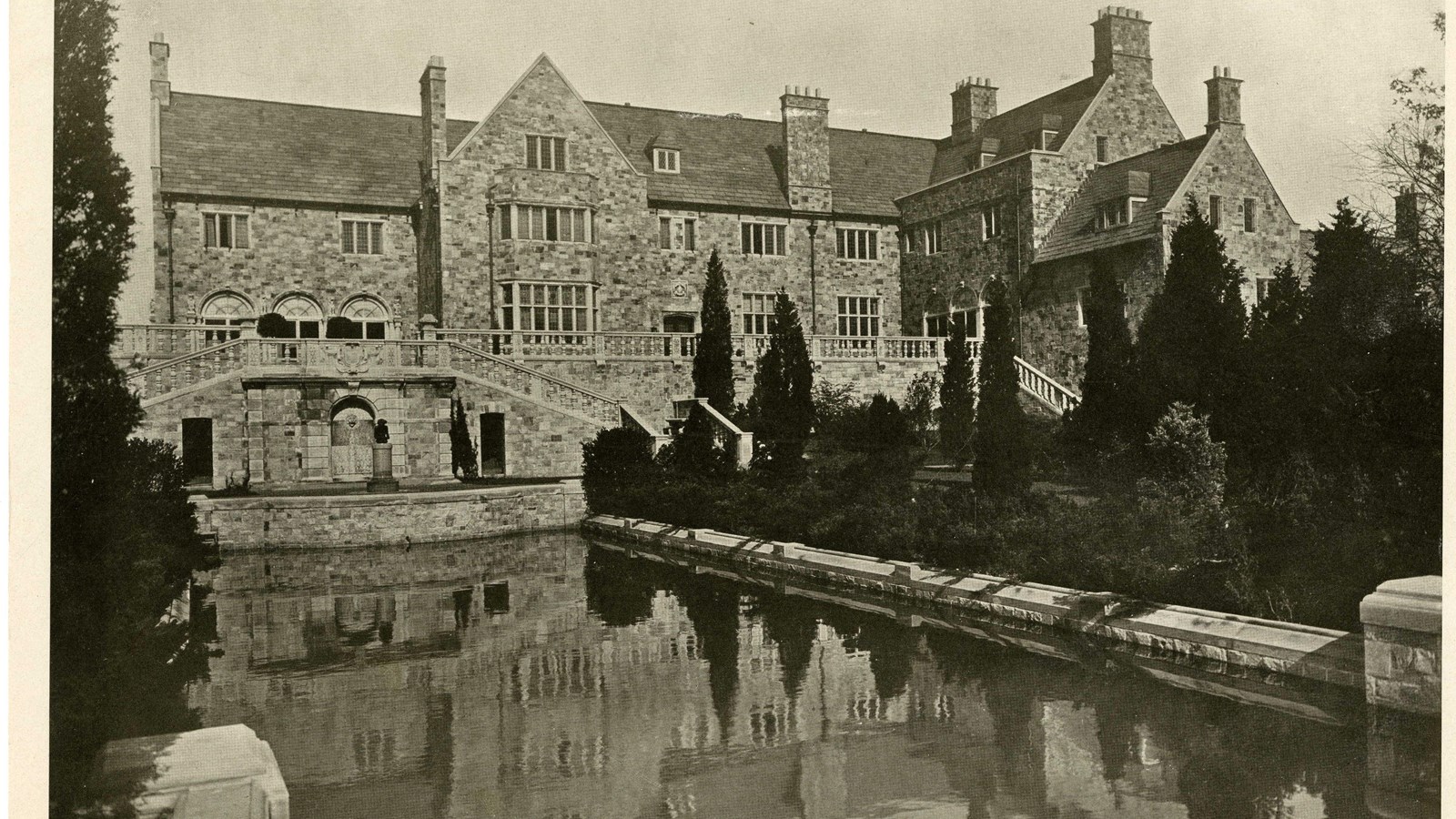 Black and white of large brick home with rectangular pool in front, trees next to it
