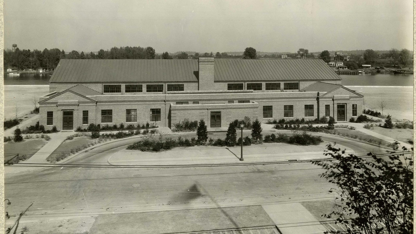 Black and white of symmetrical building with grassy area and road in front lined with shrubs