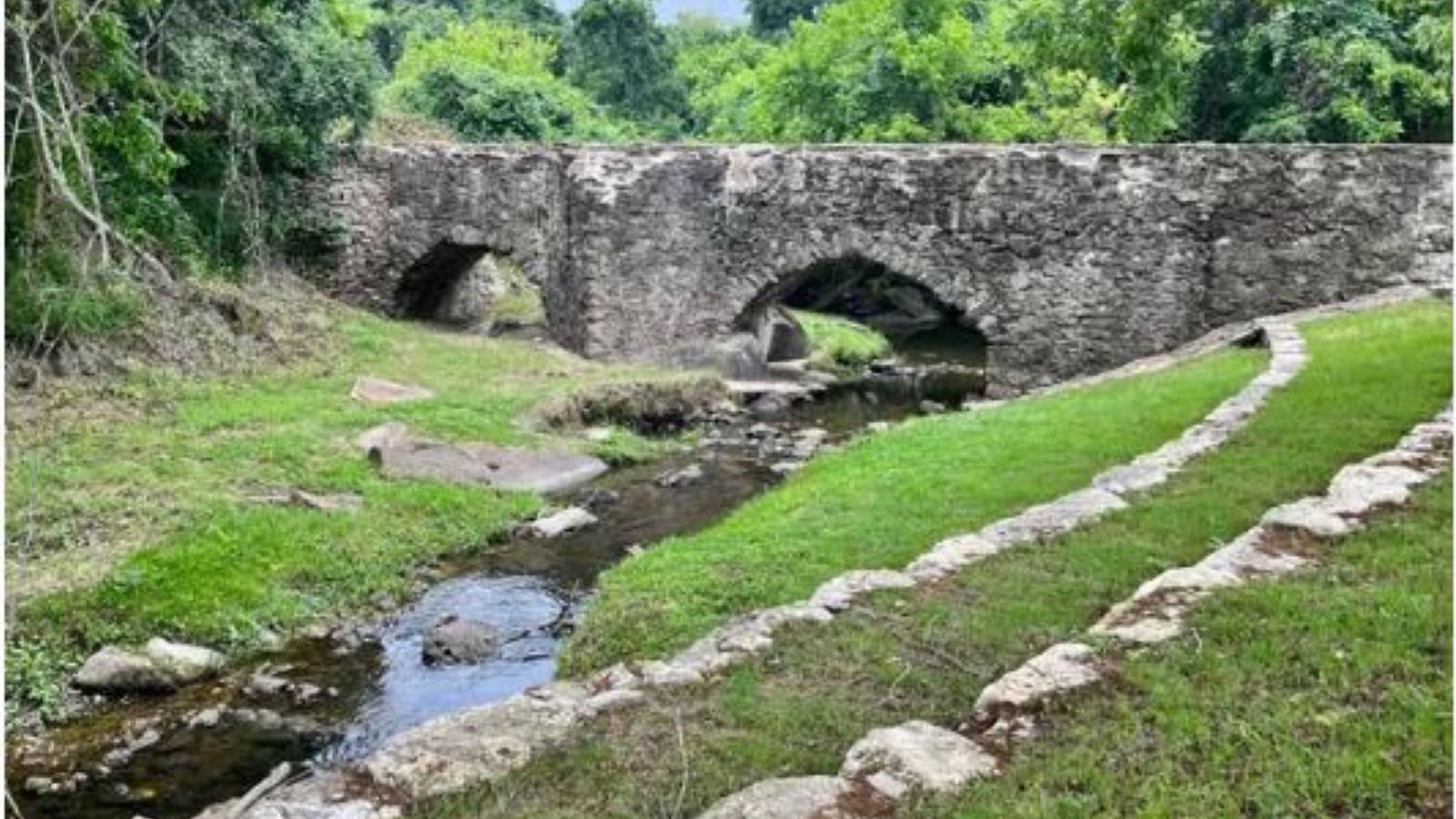 A small creek running under a stone aqueduct wall with green grass and trees on either side