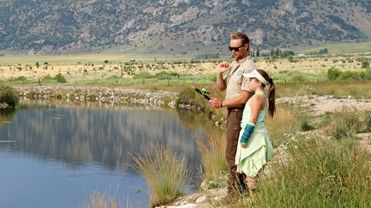 A park ranger helps a young girl fishing on the bank of the pond.