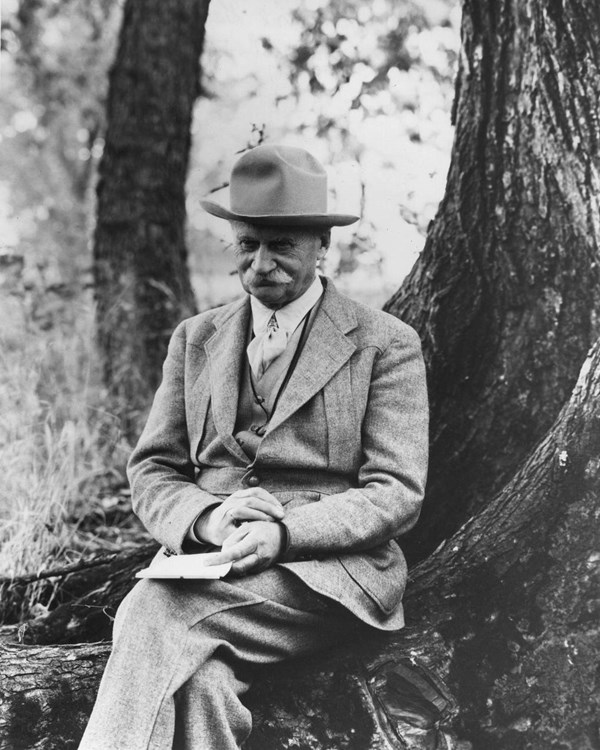Black and white photograph of a man with a white mustache wearing a suite and a hat. He is seated.