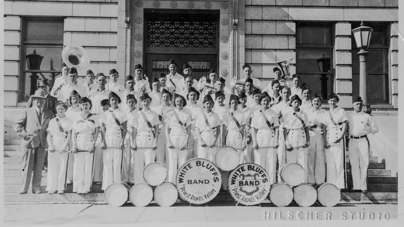 Historic photo of a school band in uniforms standing in front of a brick building