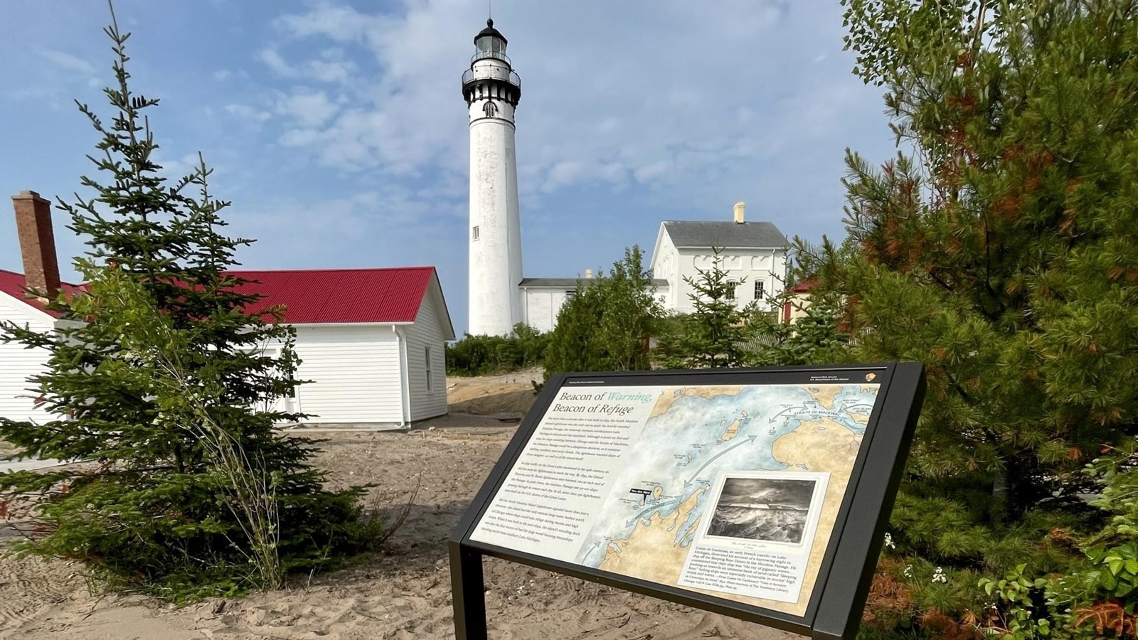 A cantilever wayside sits on a wooden platform with a white lighthouse and historic buildings behind