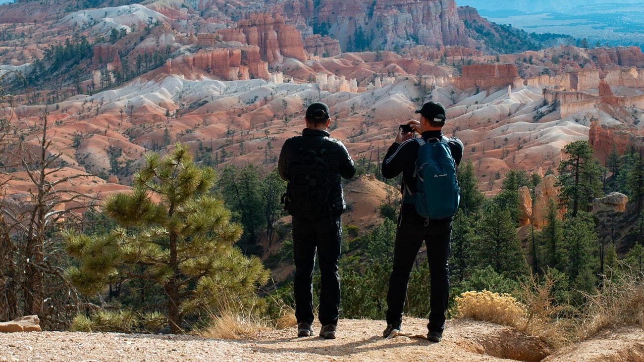 Two hikers pause along a path to view a red rock landscape of badlands and spires at a cliff\'s edge