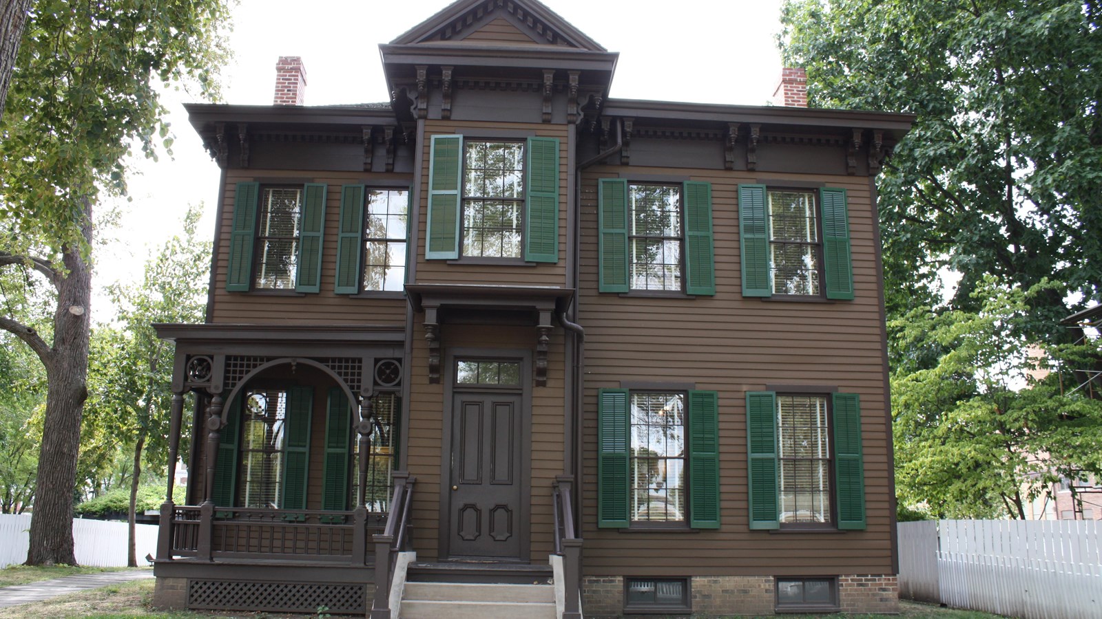 2 story dark brown house with green shutters and a small front porch left of central front door