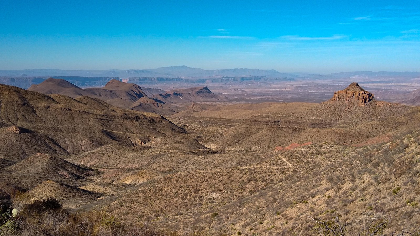 A trail descends a desert mountain slope, offering views of the surrounding mountains and plateaus.